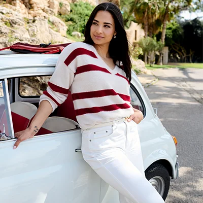 Eine Frau in einem gestreiften Pullover und einer weißen Hose lehnt an einem weißen Oldtimer.
